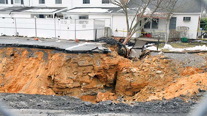 A large hole where the road has collapsed in front of a row of homes. Chuncks of rock hang on the side of the hole.
