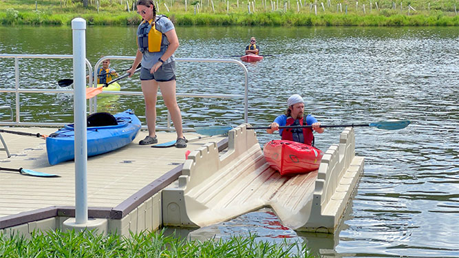 A person lowers themself onto a kayak that sits on a platform on a calm lake. There are handrails and benches on the platform.