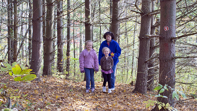 A woman and two young girls wear warm clothes and hikethrough some tall pine trees. A small, round sign and arrow on a tree