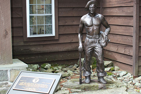 A statue of a man holding and ax stands outside a wood sided building. A plaque stands in front.