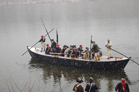 A long, open top boat floats in water with several people aboard wearing revolutionary war uniforms. Three people hold long oars