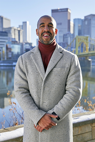 A man stands outdoors for a portrait wearing a thick winter coat. A city skyline in in the distance out of focus.