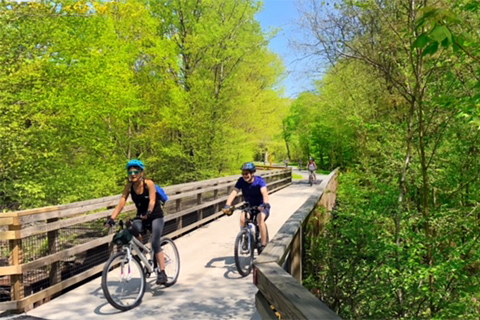 Two people ride bikes over a wooden bridge in the woods.