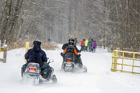 People snomobile down a snow covered trail passed an open gate.