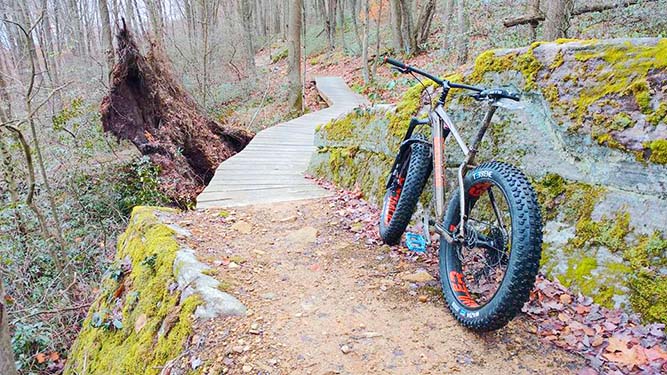 A mountain bike leans against a large boulder on a cloudy day along a dirt path which leads to a wooden boardwalk in the woods w