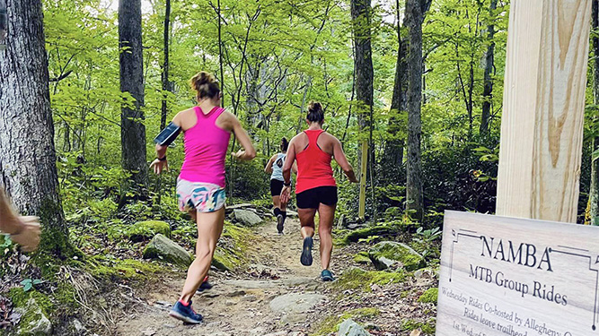  The backs of three people running on a trail past rocks and trees in a forest. A wooden sign reads: N A M B A. M T B Group Rides