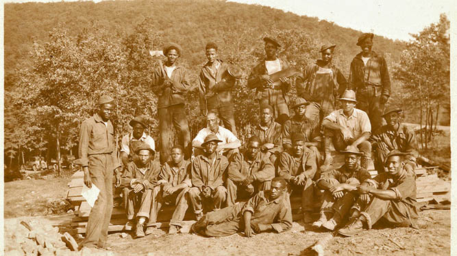 A hostoric photo of a group of men standing outdoors among trees and mountains.