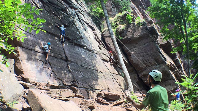 A large, flat, vertical rock face is being scaled by two people who are connected to ropes and harness. Trees and other foliage 