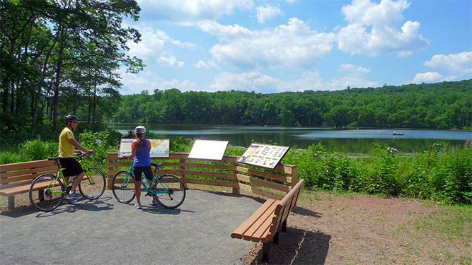 Two people stand with bicycles at an overlook of a lake with benches and informational signs.