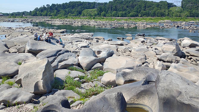 A river runs past large, sculpted boulders. A group of people sit on a boulder near the water. Trees line the far shore.