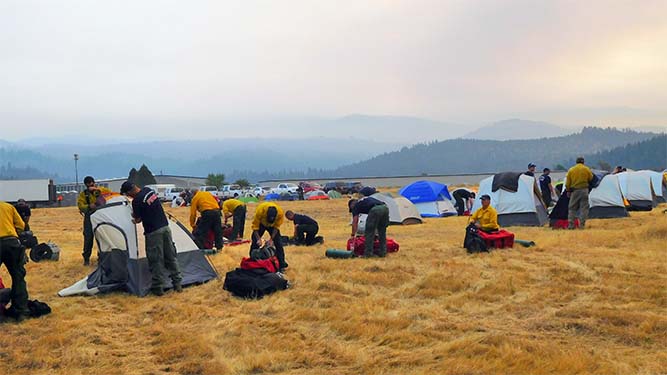 A large field of short grass in front of a wide panorama of smoke filled mountains. The field is filled with people and tents