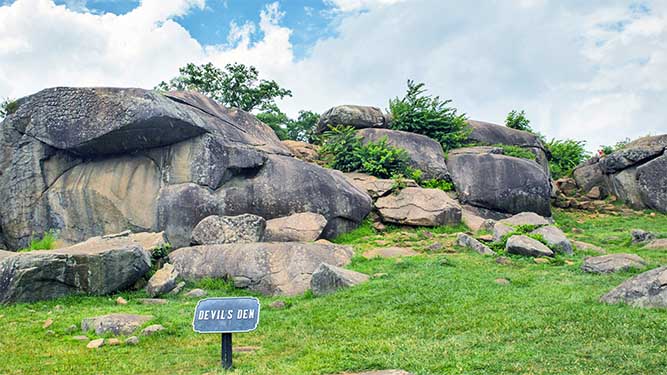 House sized boulders stick out of a small, grassy hillside with smaller ones laying on the ground. A short sign says: Devils Den