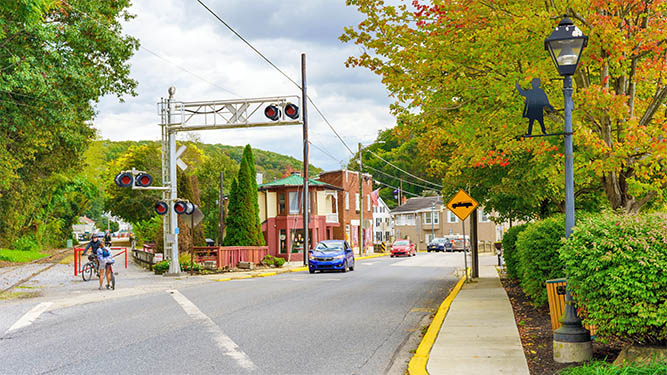 A street in a small town crosses a rail road crossing as a car drives down the street. Two people on bikes wait to cross.