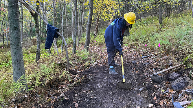 Young woman wearing hardhat uses a trail tookl with a long handle to level a section of trail.