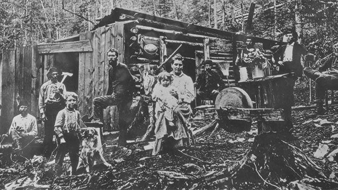 Historical photo of people posing in front of a wood hack in the woods. A woman stands with a child on her hip.