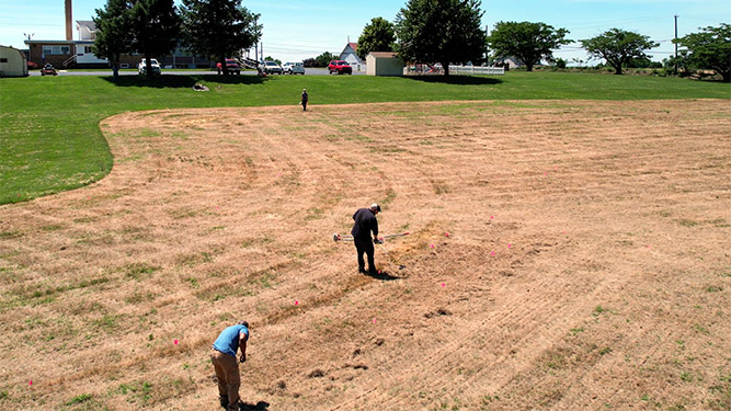 Three peopls stand seperately in a mowed field using hand tools to work the ground near a building, trees, and parking lot.
