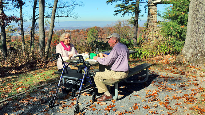 Two older adults sit on a picnic bench at a flat, gravel area in front of a vista of distant mountains.