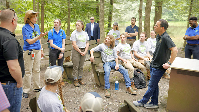 Gov  Shapiro leans against wooden podium in outdoor classroom with a group of peoplelistening to DCNR Secretary Cindy Adams Dunn