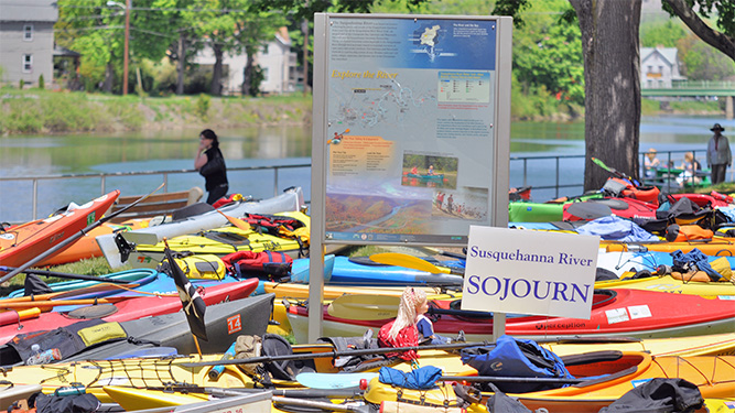 A large amount of kayaks and canoes sit in a group around a park sign. A river is in the background lined with trees and houses.