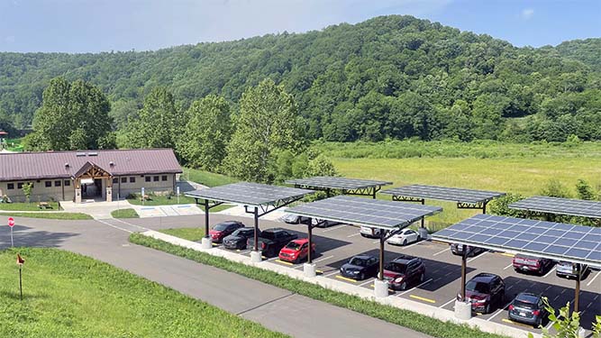 A building and parking lot with solar panels shading the parking spaces in front of a forested hill and green valley.