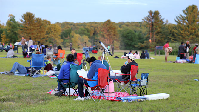 A crowd of people sit in a field in camp chairs and blanket. Some groups of people have a telescope set up.