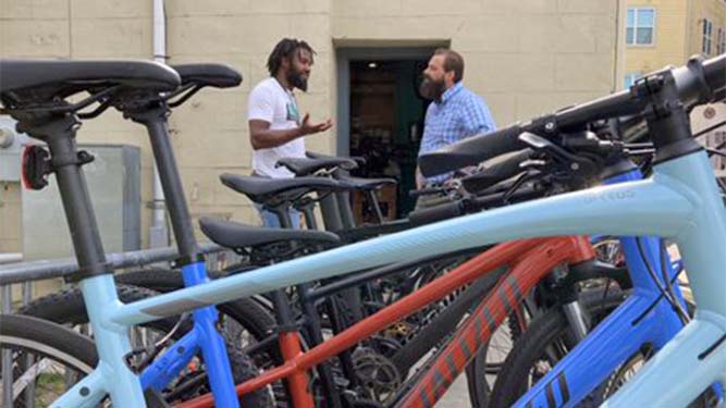 A row of bicycles stands outdoors in front of two people talking to eachother in front of a building.