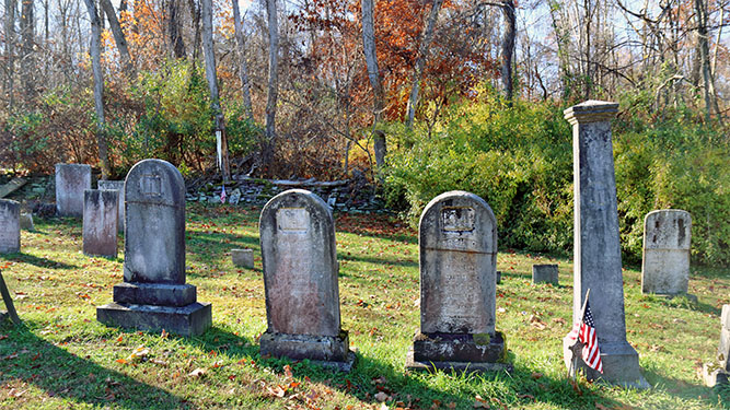 A group of headstones sit i na cemetery in front of woodlands. An American flag sits at the base of one of the headstones.