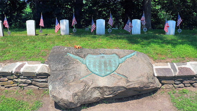 A metal plaque set into a large rock in front of a small rock wall and a row of small, white headstones with American flags