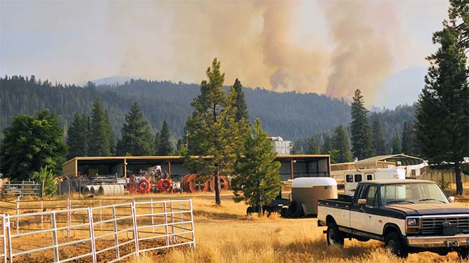 Smoke rises in a plume and fills the air above a forested mountain, a garage with vehicles and equipment, and a pick up truck.