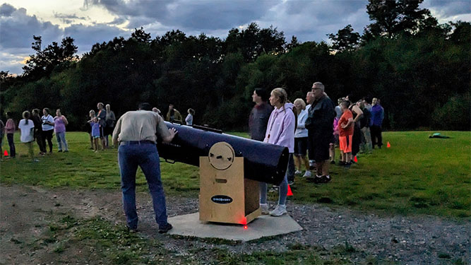 People stand outdoors during dusk in a field in front of trees, standing around a tube shaped telescope