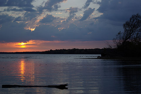 A large lake is rippled as the sunset dips behind clouds. Forested shores can be seen in the distance.