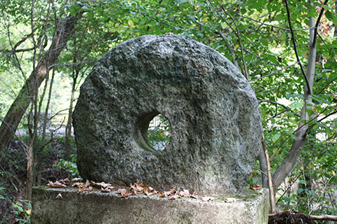 A stone griding wheel still uncut from the original block it was carved from stands outdoors in a forest. 