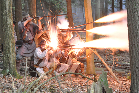 A group of peopls in frenc hand indian war reenactment uniforms fire muskets in the woods with long, bright sparks and smoke.