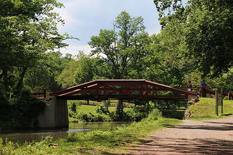 A wooden truss bridge spans a canal amongst trees and plants, and a walking path