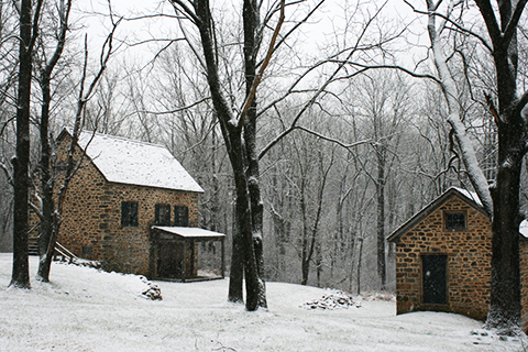 Two stone buildings stand outside in the woods with light snow covering everything