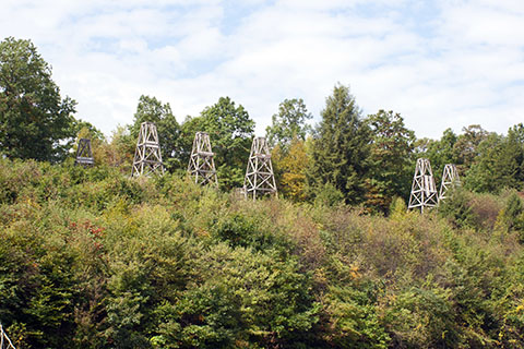 Wooden oil dericks stand on a brushy and wooded hillside.