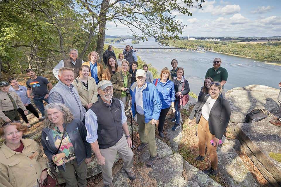 A large group of people pose for a photo at an overlook over a large river. People stand on the rocky outcrop with trees to the 