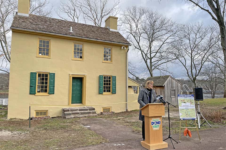 A woman stads and speaks at a podium outdoors in front of a historic house.