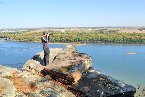 A man holding binoculars stands on a rocky outcrop above a river with trees, fields, and roads in the distance.